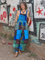 Woman wearing wide-leg blue and green patchwork dungarees with printed panels and a front pocket, leaning against a graffiti-covered brick wall